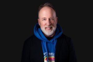 Portrait of a smiling man with a beard in a blue hoodie and black jacket.