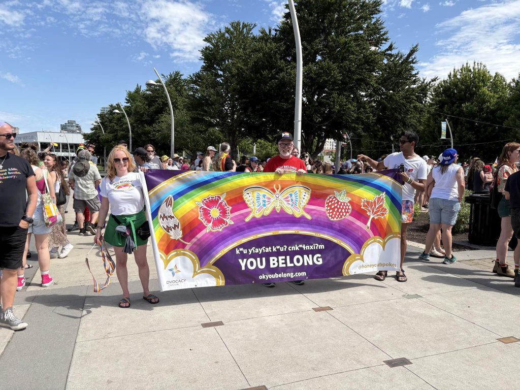 A group of people at a Pride event holding a colorful banner that reads “YOU BELONG” with rainbow and floral designs, surrounded by a lively crowd on a sunny day.