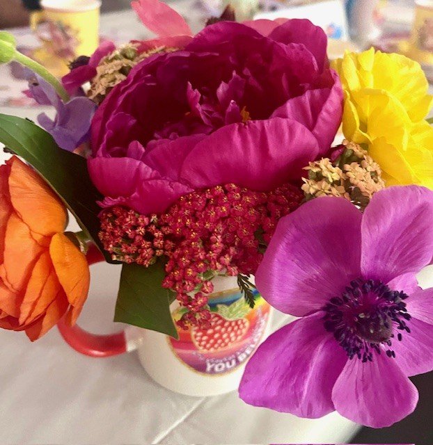A colorful bouquet of flowers, including purple anemone, orange ranunculus, pink peony, yellow ranunculus, and red yarrow, arranged in a mug that says 