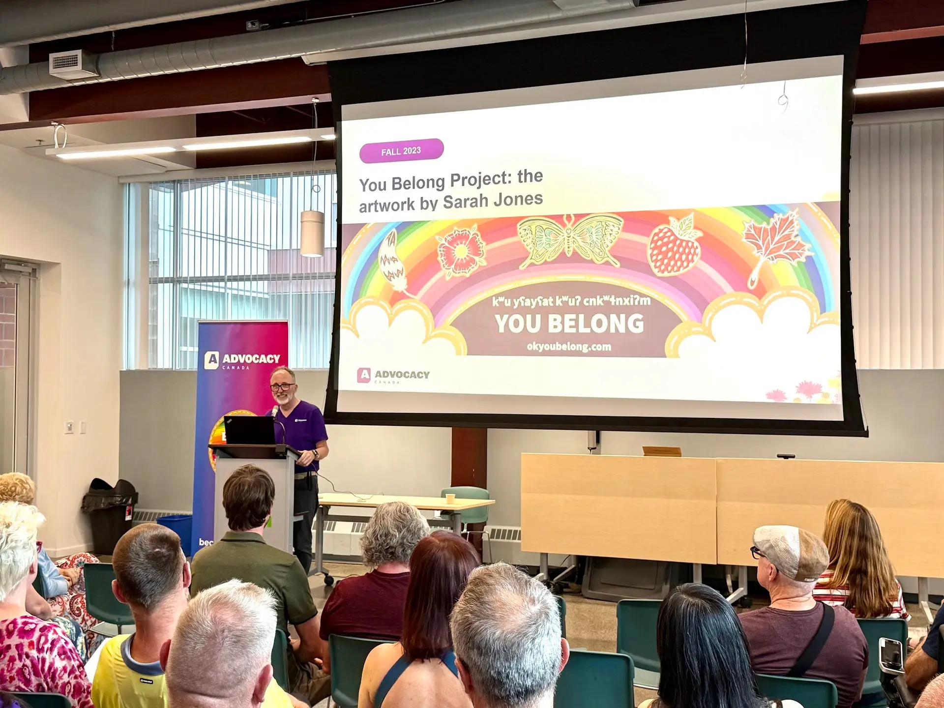 A man stands at a podium presenting to a seated audience in a community room. He wears glasses and a purple shirt. Behind him, a large projector screen displays a slide titled "You Belong Project: the artwork by Sarah Jones," dated Fall 2023. The slide features colorful rainbow artwork with illustrations of a feather, flower, butterfly, strawberry, and maple leaf, along with Syilx language text and the words "YOU BELONG" and "okyoubelong.com." An Advocacy Canada banner stands beside the podium. Audience members are seated in rows of green chairs facing the screen.